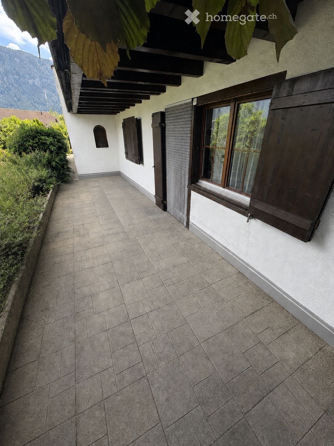 A narrow, tiled corridor with a white wall on one side and a wooden-framed window on the other, leading to a covered outdoor area with a view of mountains.