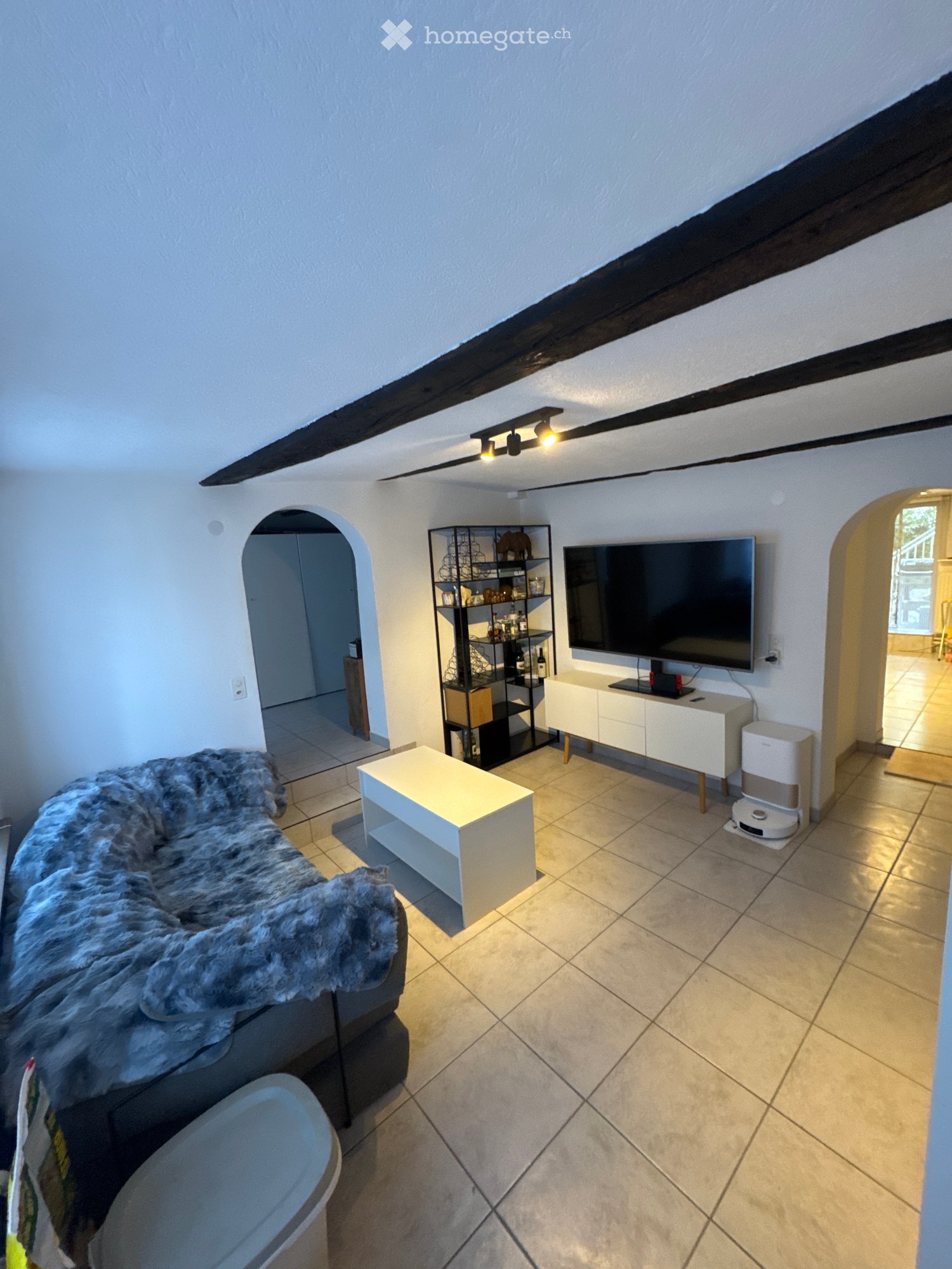 Modern living room with a grey sofa, white coffee table, TV stand, shelf, and floor lamp. The room features exposed wooden beams on the ceiling and tiled flooring.
