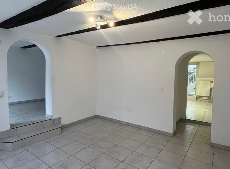 Empty room with white walls, tiled floor, and two arched doorways leading to other spaces. The room has a raised platform on one side and a ceiling with exposed wooden beams and a central light fixture.