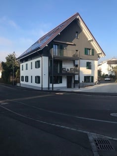 Modern two-story house with white exterior, dark green shutters, and a balcony on the upper floor. Solar panels are installed on the roof.