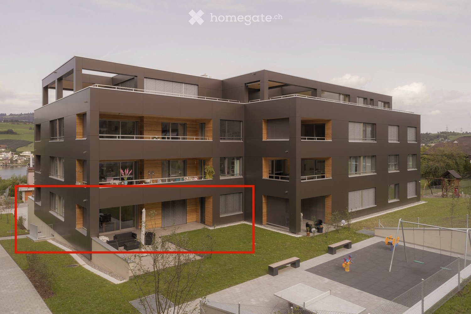 Modern multi-story residential building with balconies, large windows, and a landscaped courtyard featuring benches, a swing set, and a patio area.