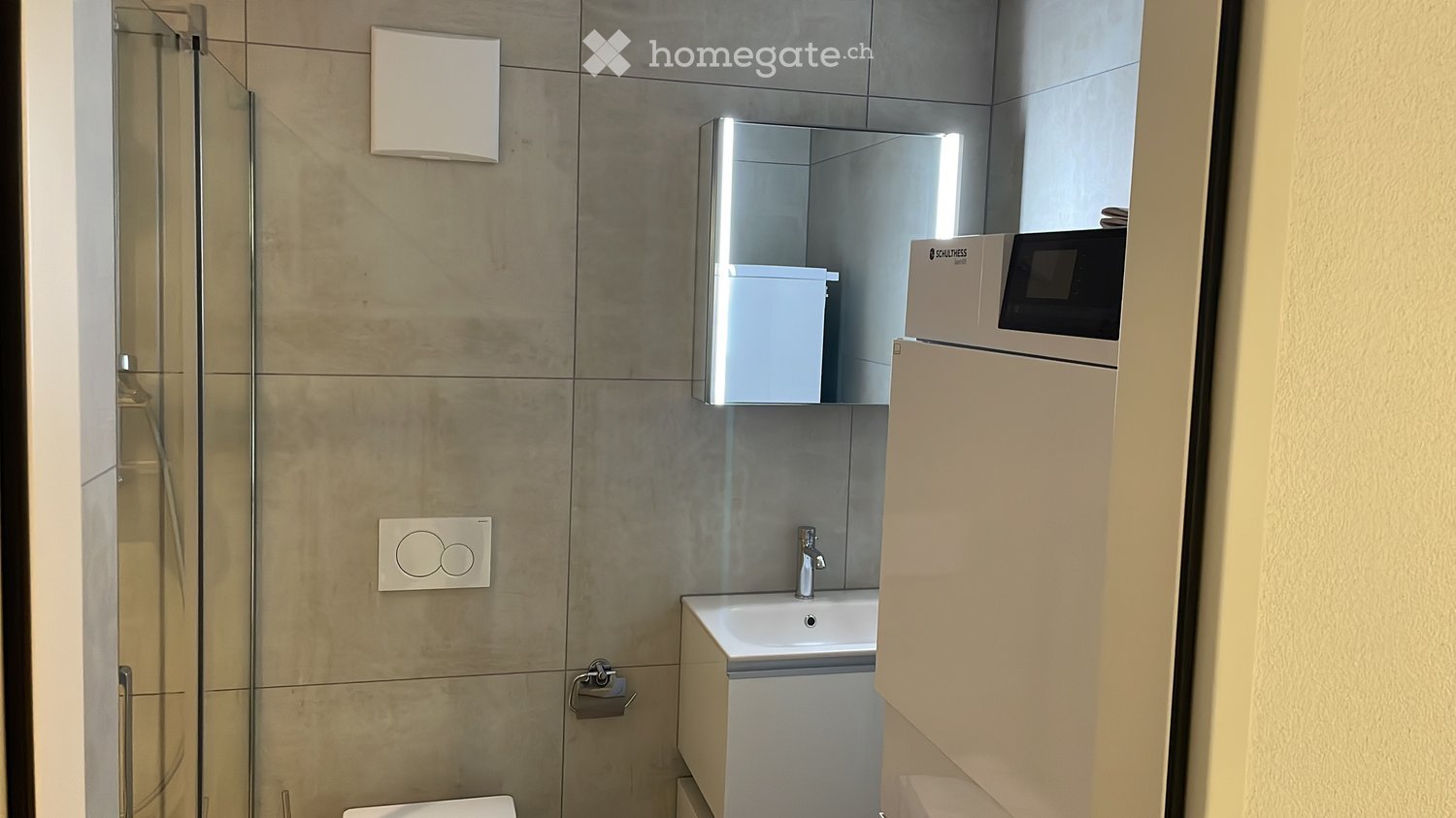 Modern bathroom with a glass-enclosed shower, a white sink with a chrome faucet, a mirror cabinet, and a toilet. The walls are tiled in a neutral color, and there is a small cabinet next to the sink.