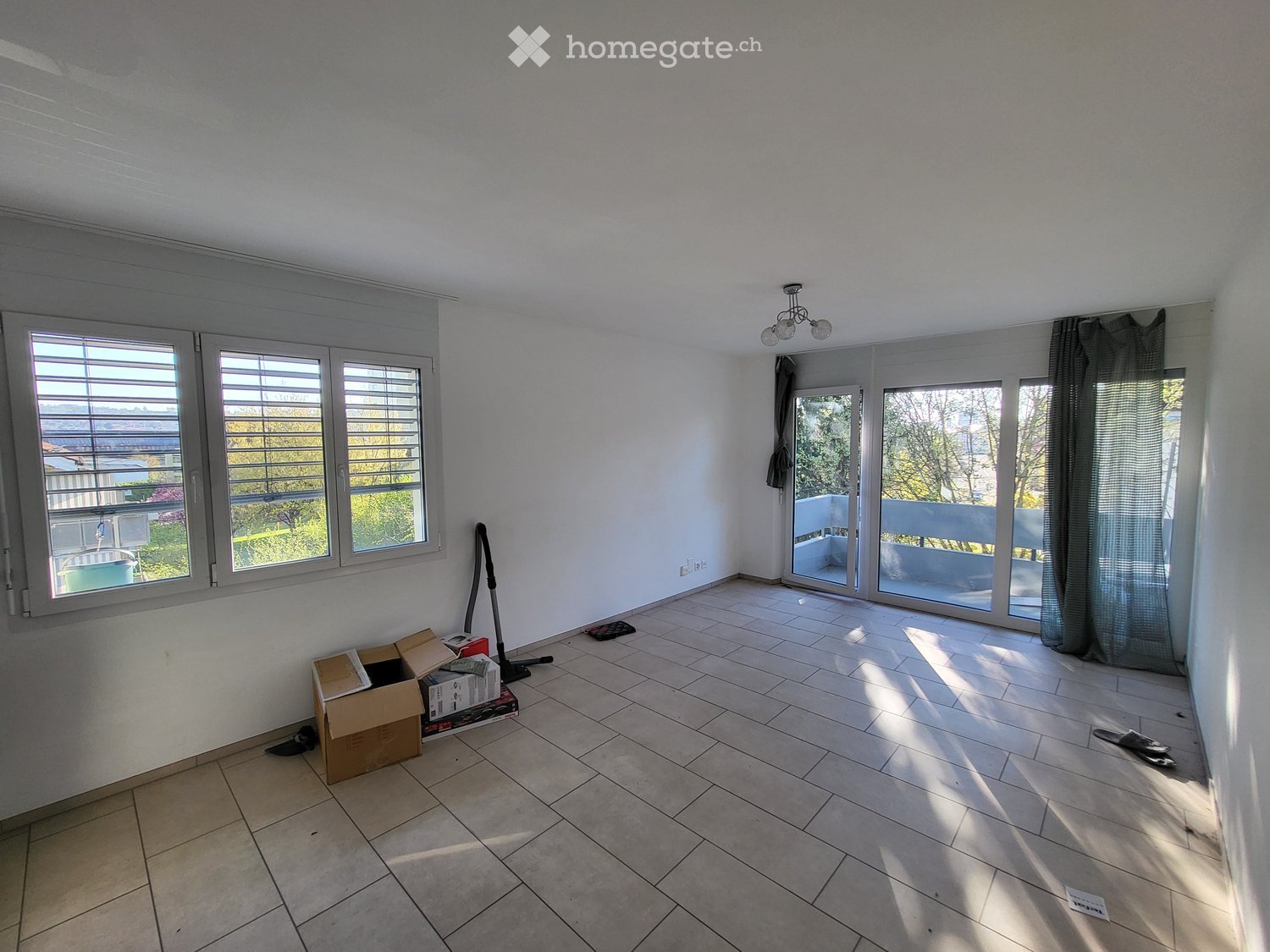 Empty room with tiled floor, large windows, sliding glass doors leading to a balcony, and some scattered items like a vacuum cleaner and boxes.