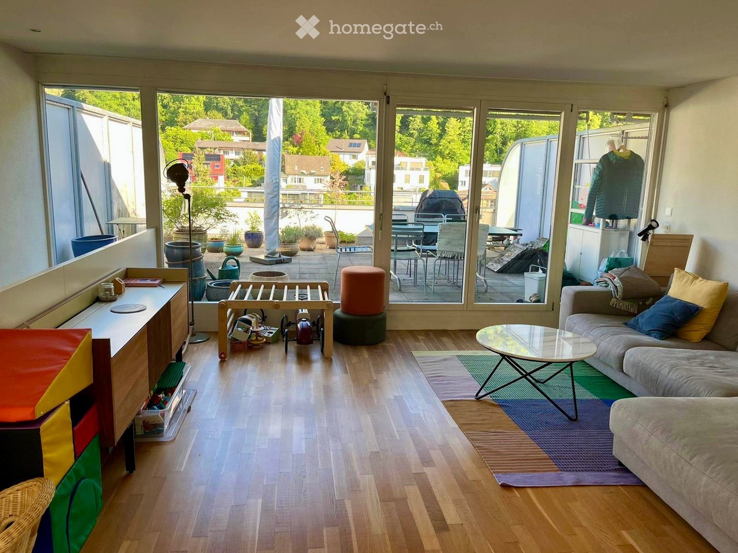 Bright living room with wooden flooring, large windows opening to a balcony, sofa, coffee table, and various decorative items.