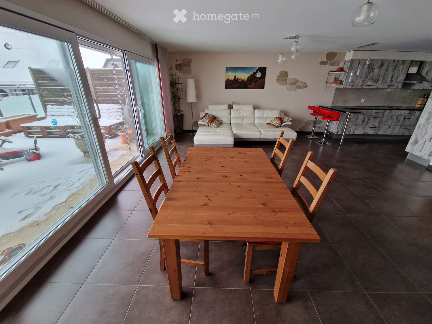 Bright living and dining area with large glass doors leading to a snowy outdoor space, featuring a white sectional sofa, wooden dining table with chairs, red bar stools, and modern lighting.