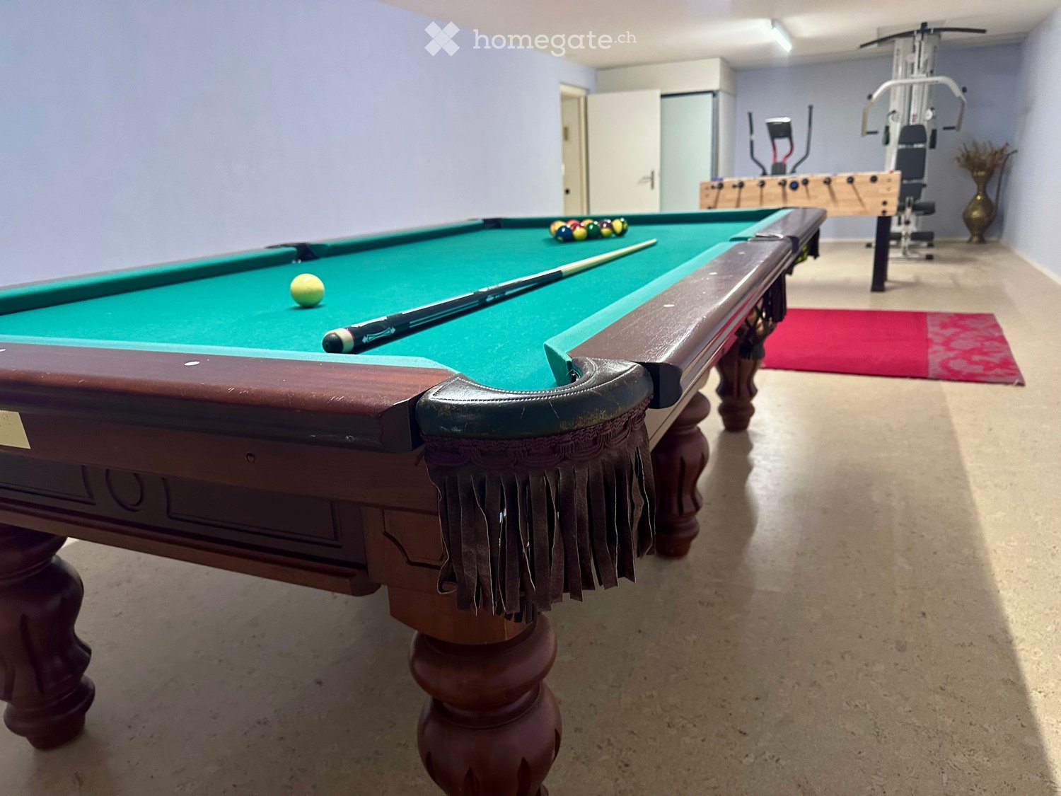 Billiard room with wooden pool table, green felt, billiard balls, and cue stick. The room has light-colored walls, a white ceiling, a red carpet, and other sports equipment.