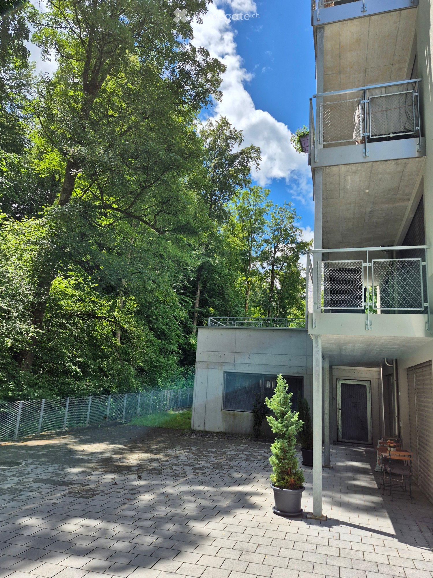 Modern outdoor area with a paved patio, small potted tree, and a small seating area with a table and chairs. The area is surrounded by greenery and has a modern building with balconies in the background.