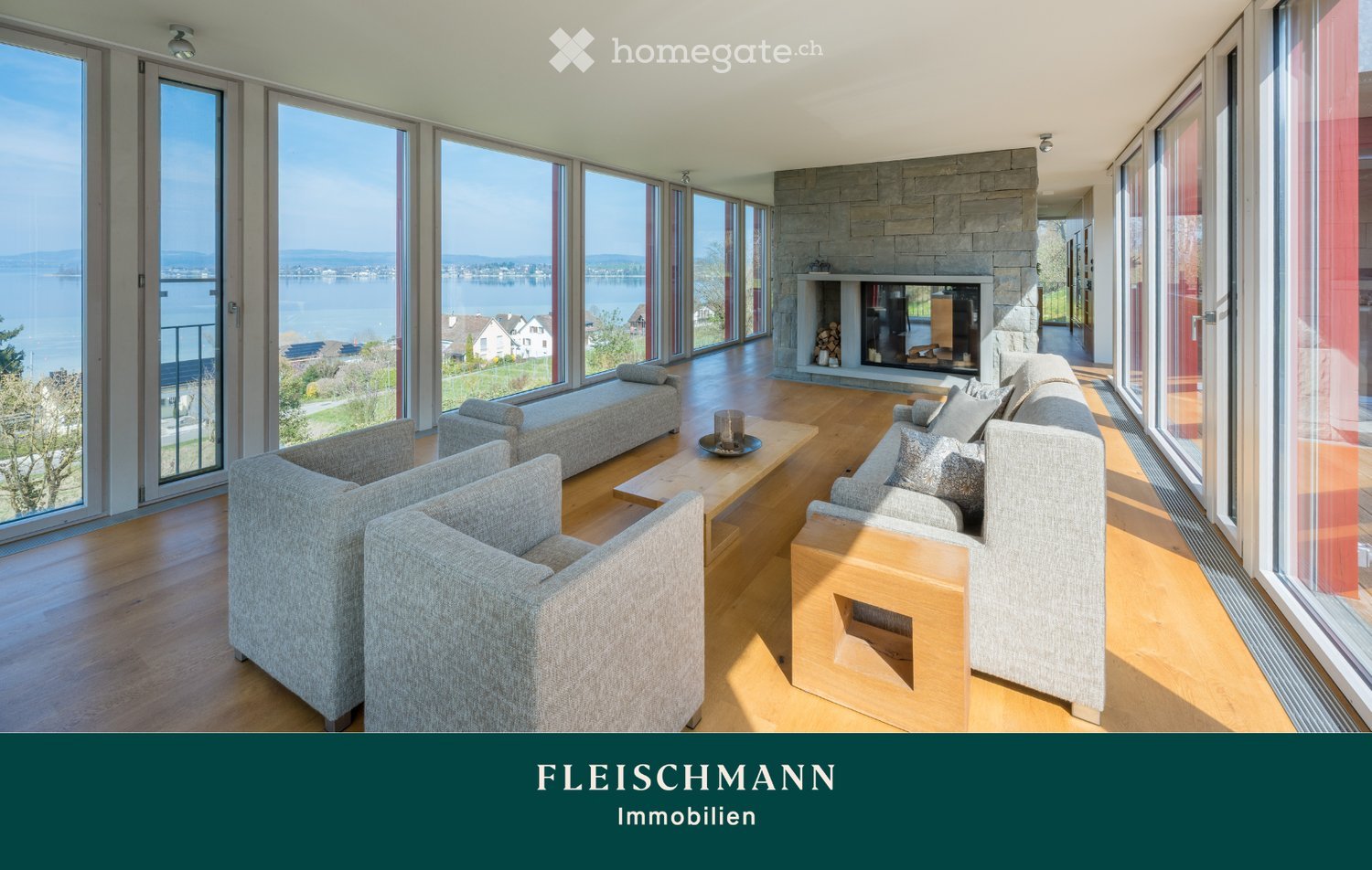 Modern living room with large windows offering a scenic lake view, featuring a grey sofa, coffee table, armchairs, ottoman, and a stone fireplace.