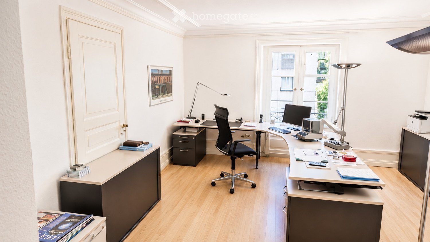 Modern office space with light wood flooring, white walls, and large windows. Features a black office chair, a desk with a computer, a printer, and a lamp, along with a cabinet and a picture frame on the wall.