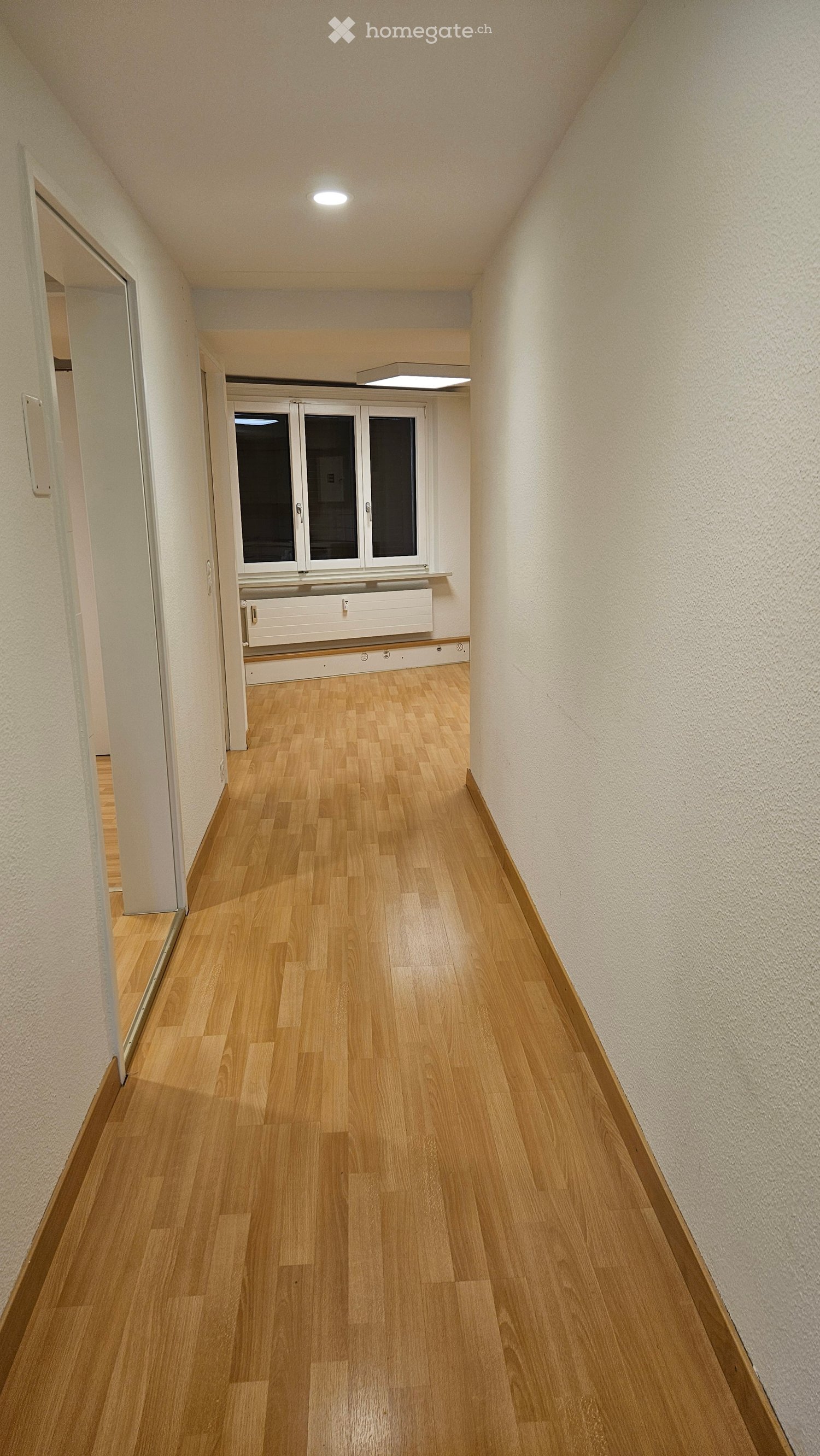 A clean, well-lit hallway with wooden flooring, white walls, and a large window at the end. The ceiling has recessed lighting, and there is a radiator beneath the window.