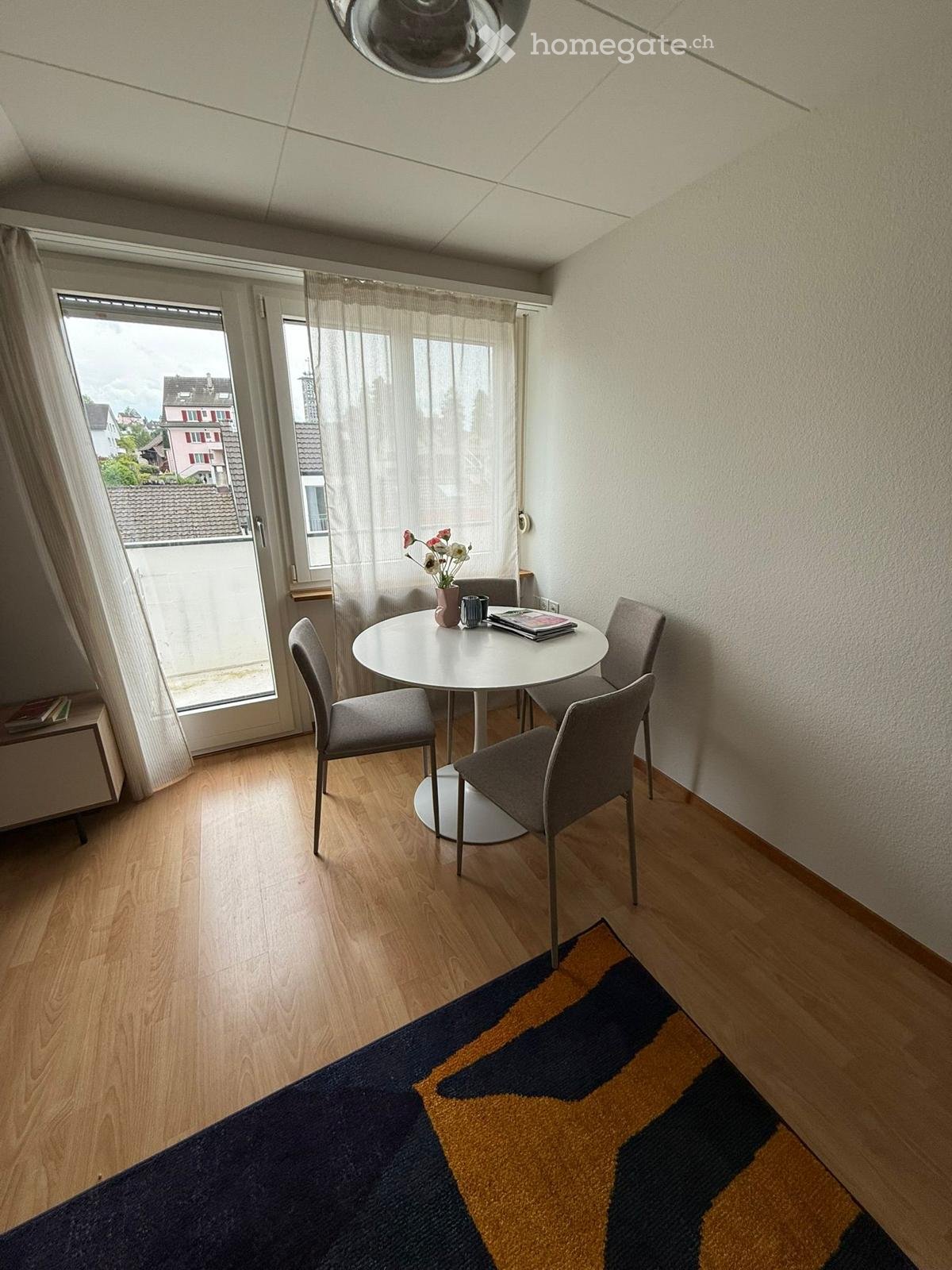 A small, modern dining area with a round white table, four grey chairs, a vase with flowers, a cup, and books on the table. The room has wooden flooring, a patterned rug, and large windows with sheer curtains.