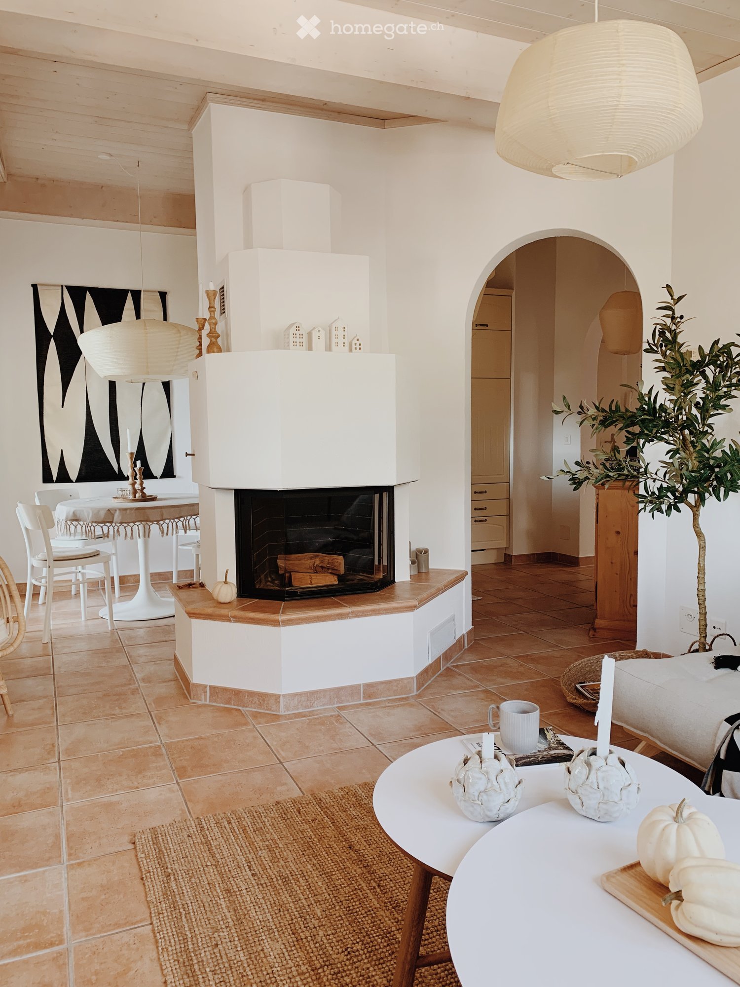 Modern living room with a fireplace, white walls, wooden ceiling beams, and a mix of contemporary and rustic decor including a sofa, coffee table, and decorative pumpkins.