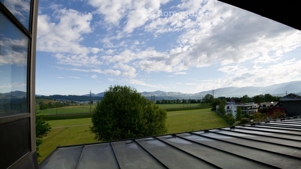 Balcony with a view of a green field, trees, and distant mountains under a partly cloudy sky.