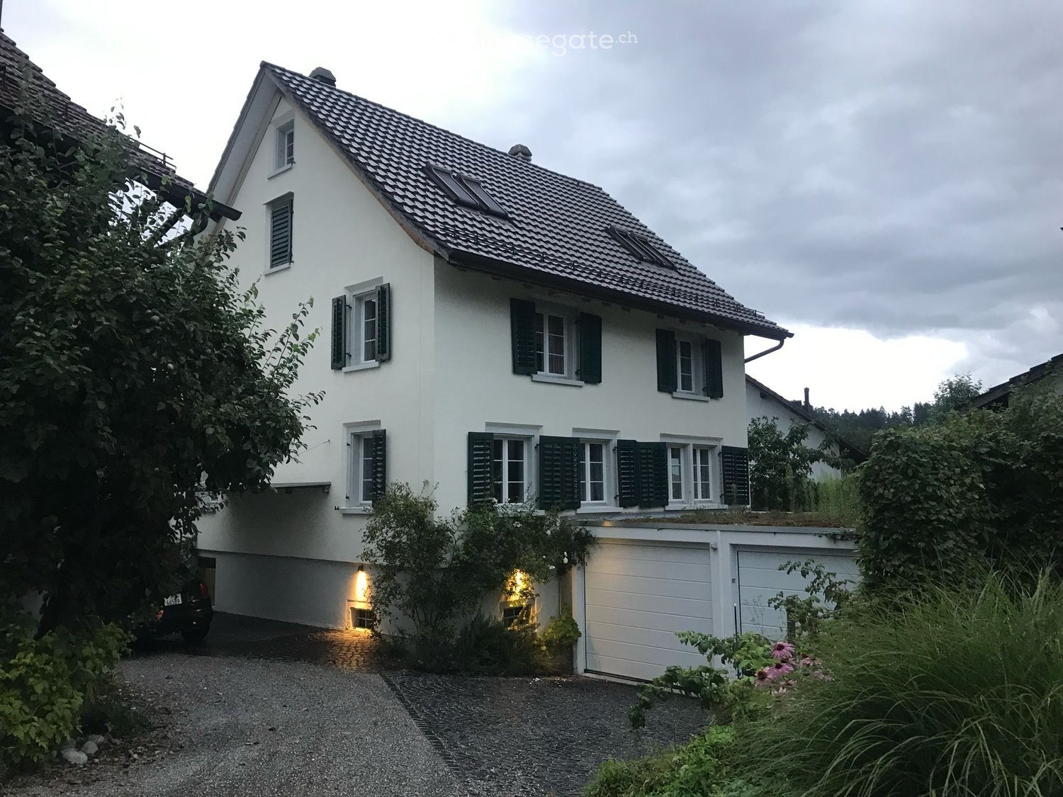 Two-story white house with a tiled roof, multiple windows with shutters, and a garage. The house is surrounded by greenery and has a gravel driveway.