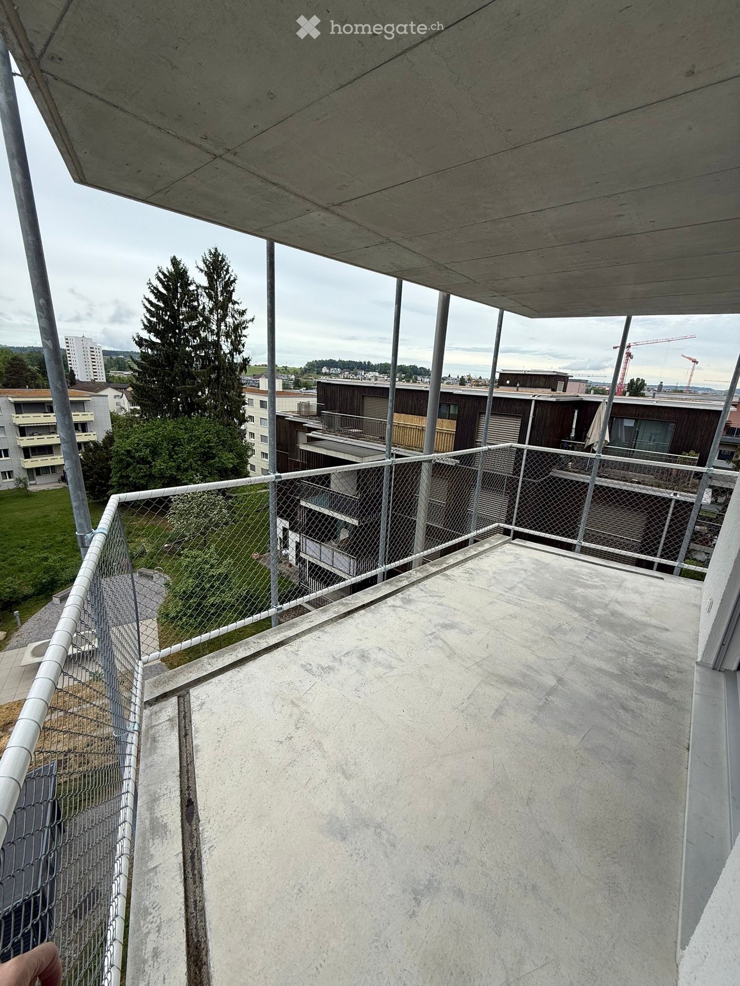Balcony / Terrace. Concrete balcony with metal railing, overlooking a residential area with buildings, trees, and a grassy lawn.