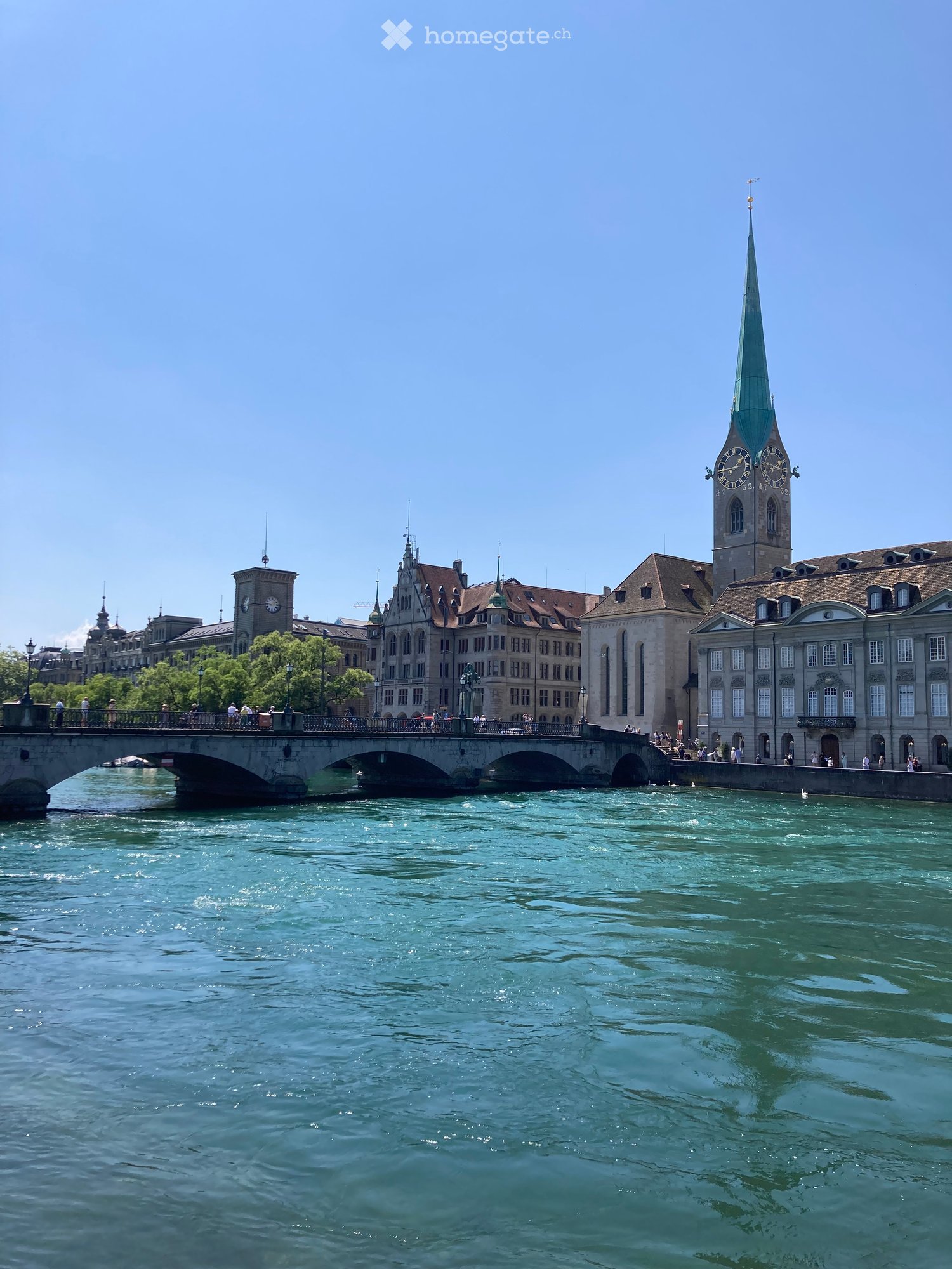 A scenic view of a river with a bridge and historic buildings, including a tall clock tower with a spire, on the opposite bank.