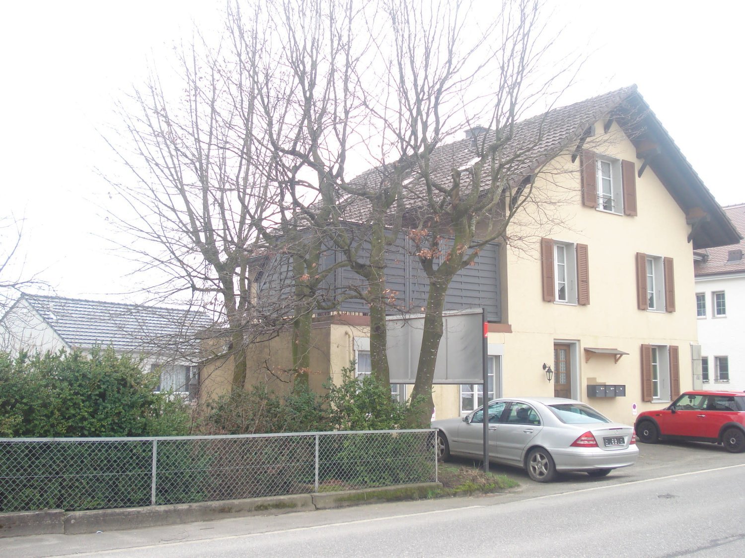 Two-story house with yellow facade, multiple windows, and a small balcony. There are two parked cars in front of the house, and a fence lines the front yard.