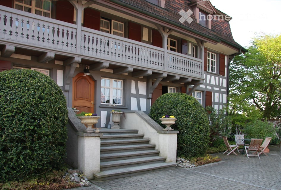 Two-story house with a balcony, wooden door, and well-maintained garden with chairs and tables.