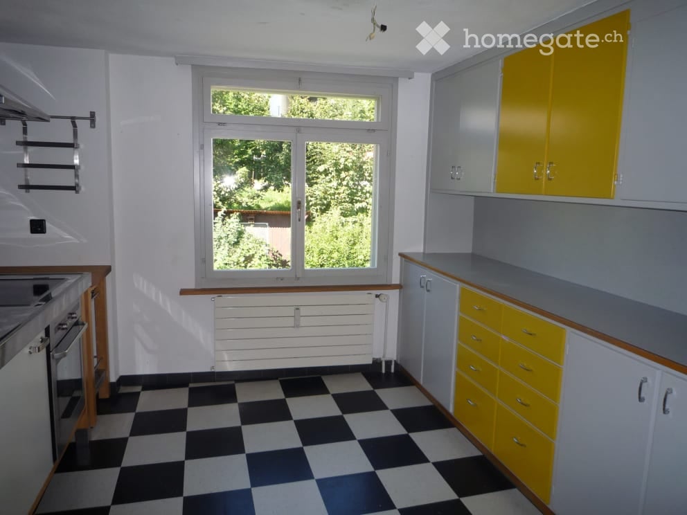 Bright kitchen with checkered black and white flooring, white walls, and yellow cabinets. Features a window with a view of greenery, a sink, and an oven.
