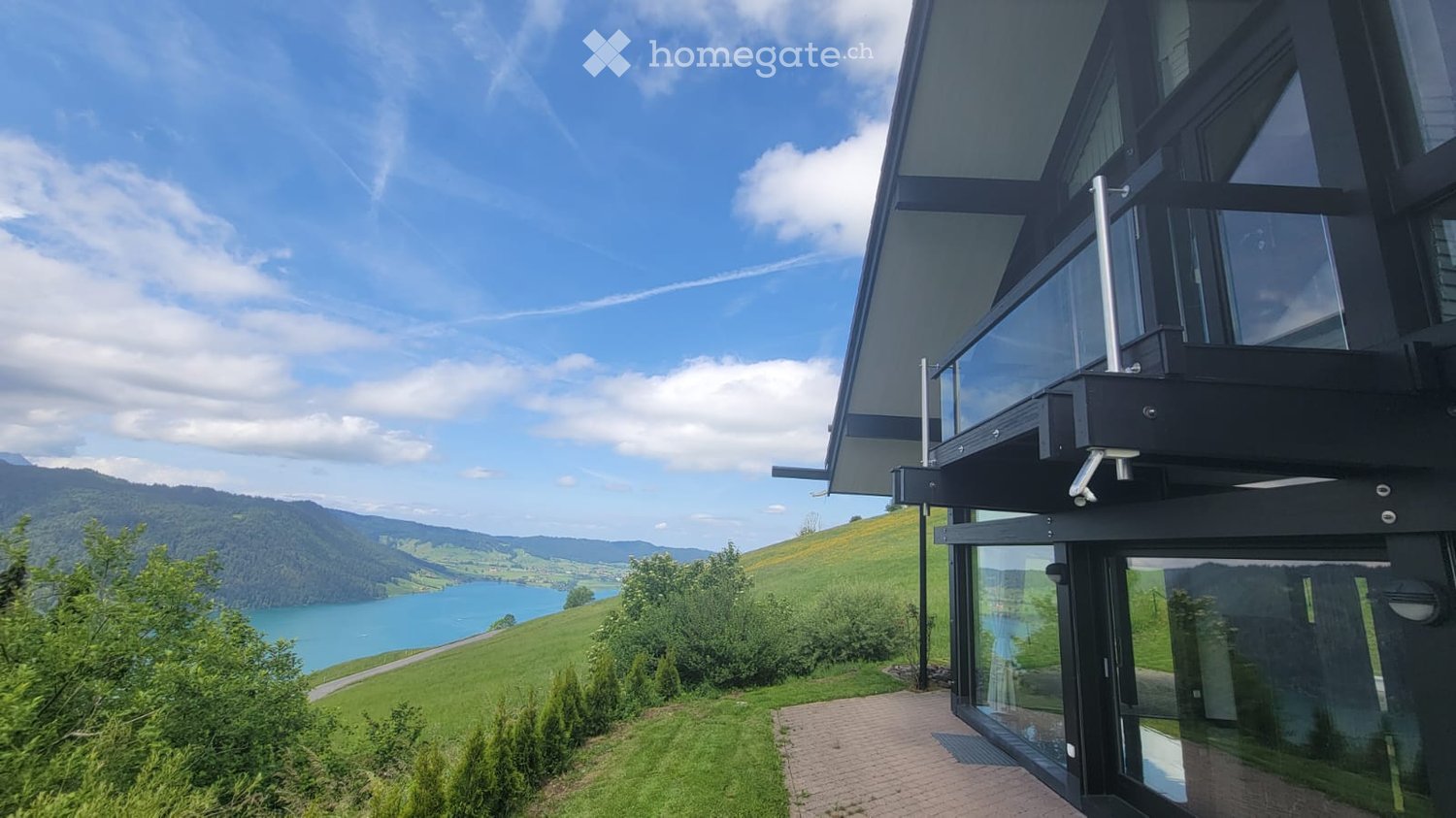 Modern two-story house with large glass windows and a balcony overlooking a scenic lake and green hills.