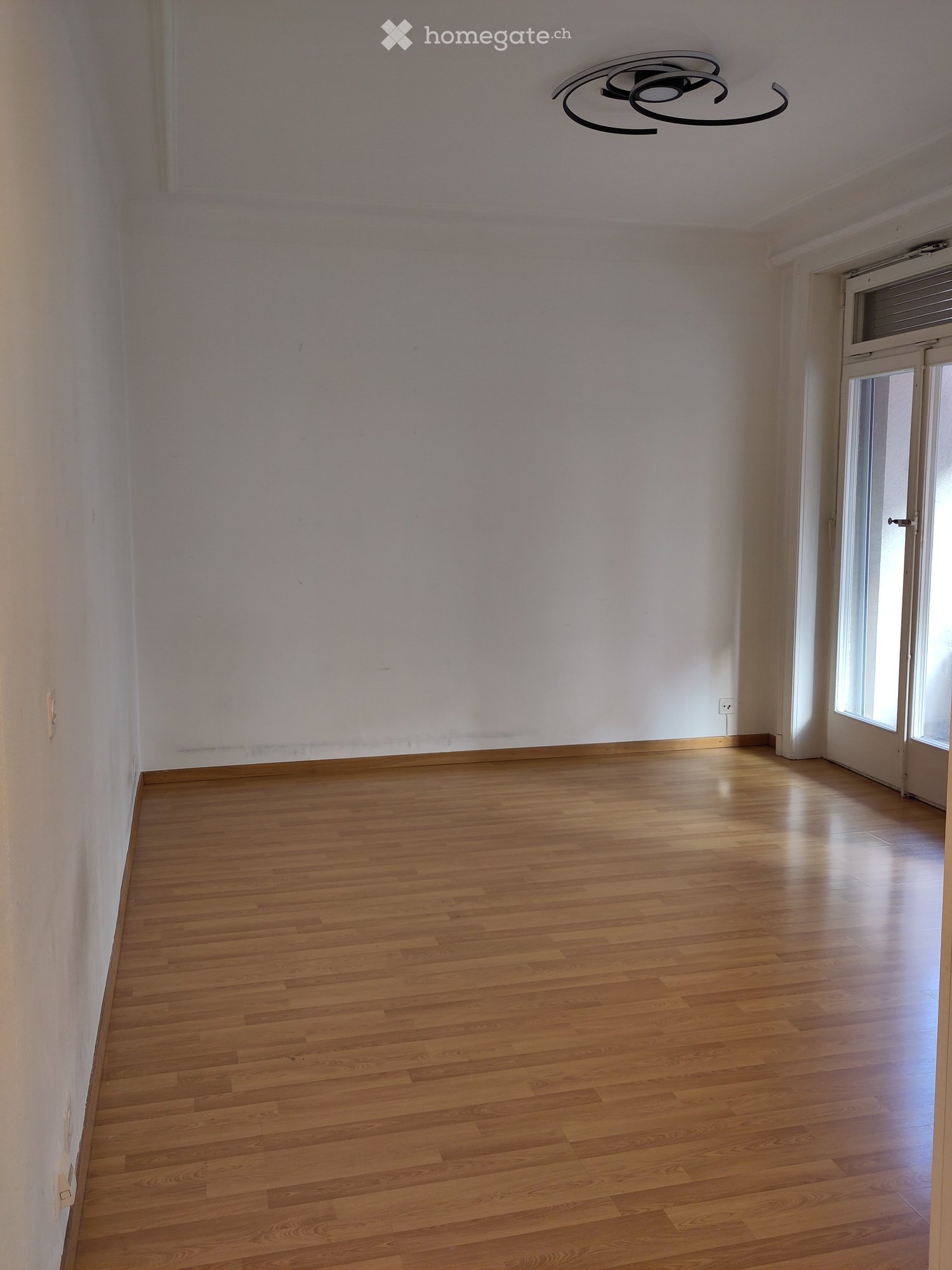 Empty room with wooden flooring, white walls, and a modern ceiling light fixture. Features a large window with blinds and a door leading to another space.