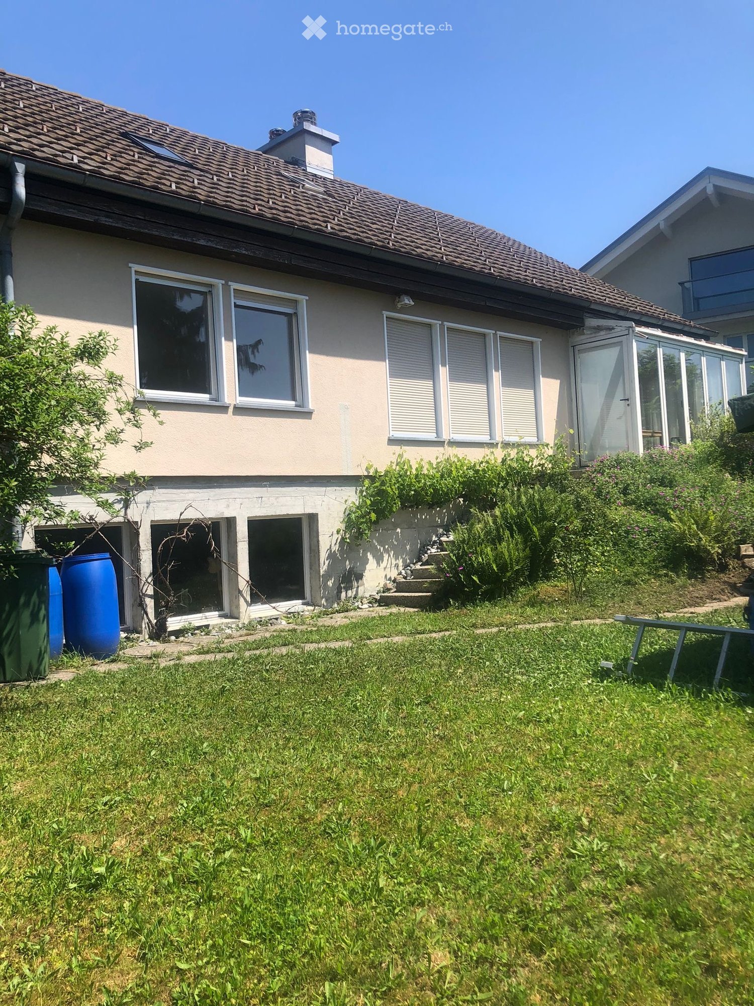 Two-story house with beige walls, tiled roof, multiple windows, and a small garden area with grass and plants.