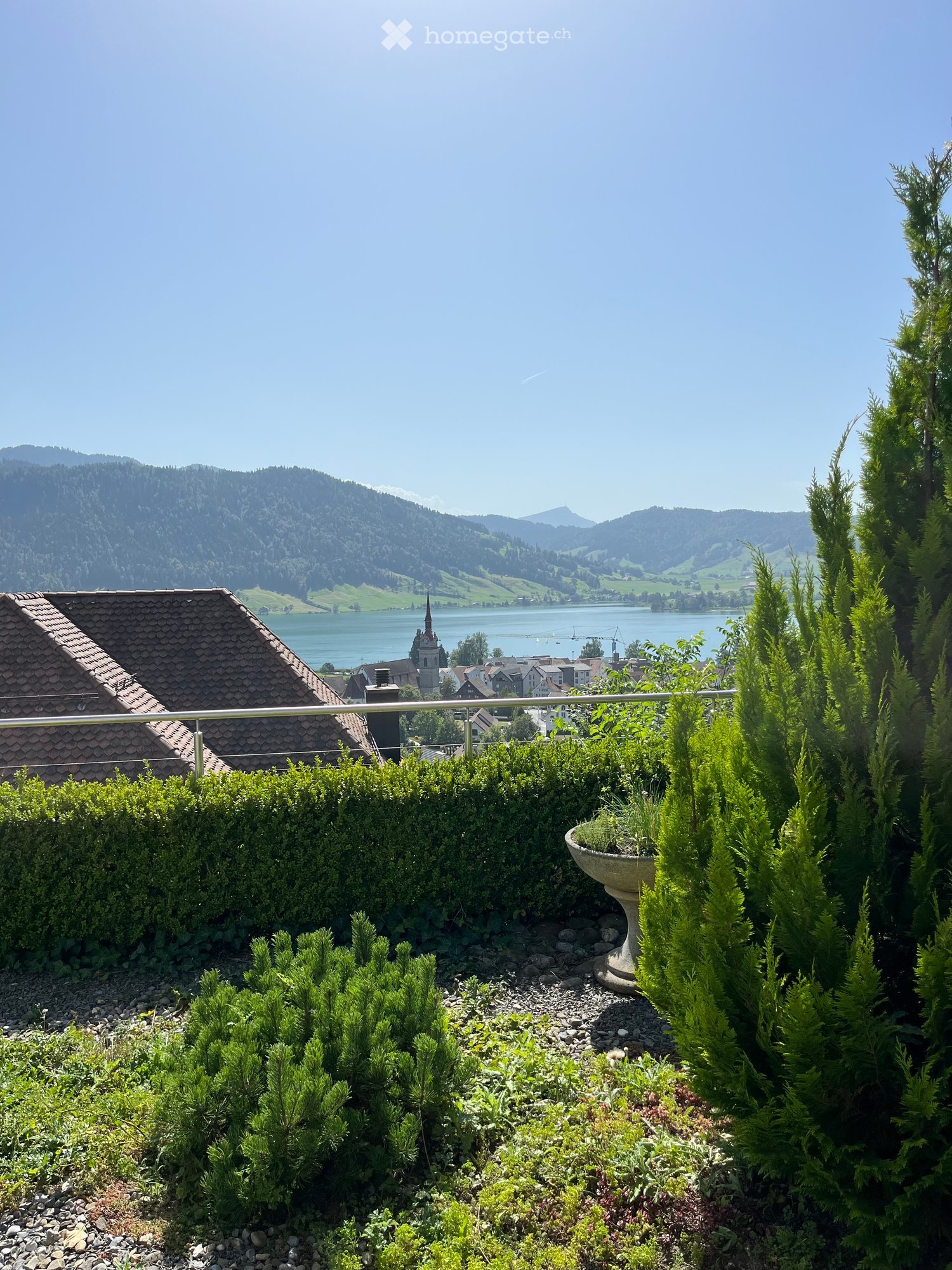 A well-maintained garden with various shrubs and trees, featuring a metal railing and a view of a lake and mountains in the background.