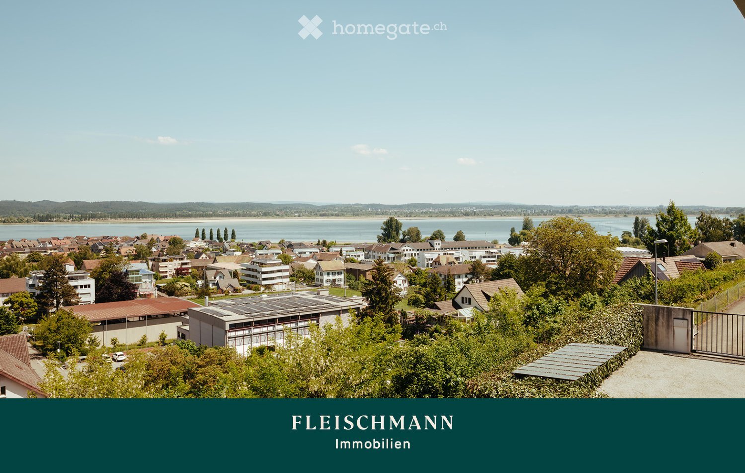 A panoramic view of a town with houses, trees, and a body of water in the background, featuring a clear blue sky and distant hills.