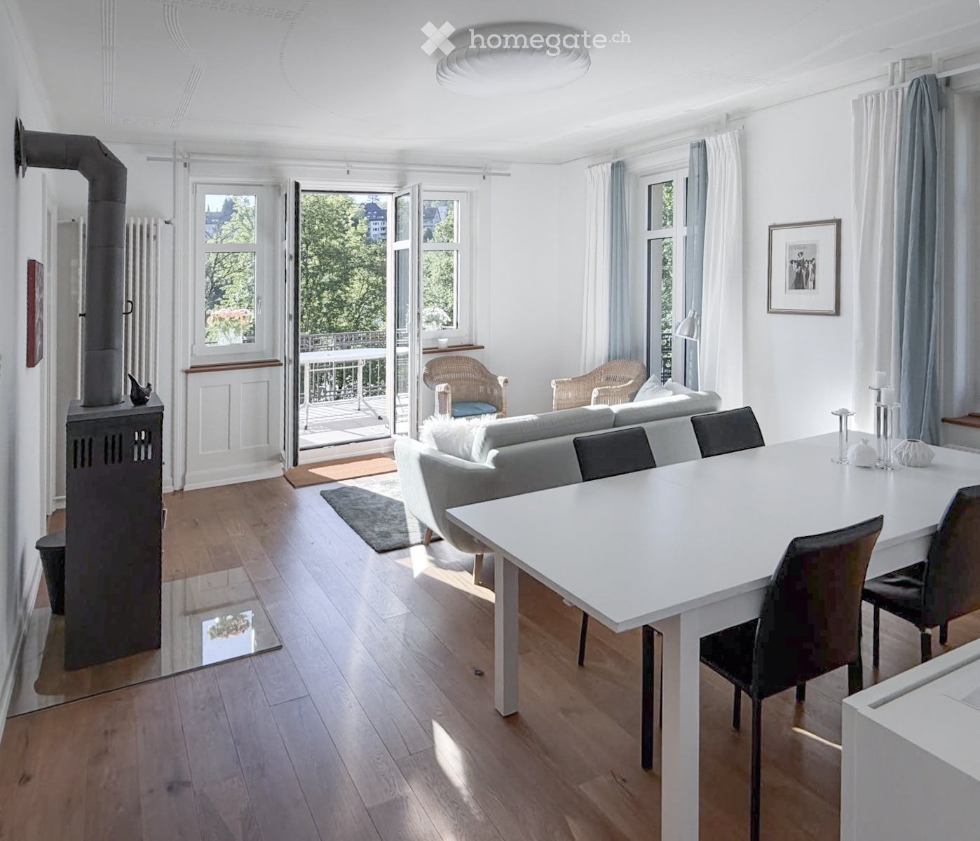 Bright living and dining area with wooden flooring, white walls, large windows, and a balcony. Features a grey sofa, dining table with chairs, wicker chairs, and a table lamp.