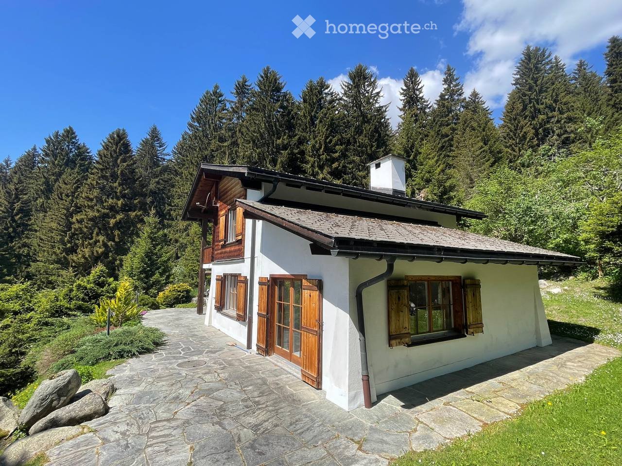 A single-story house with white walls, wooden shutters, and a stone pathway leading to the entrance, surrounded by tall trees and greenery.