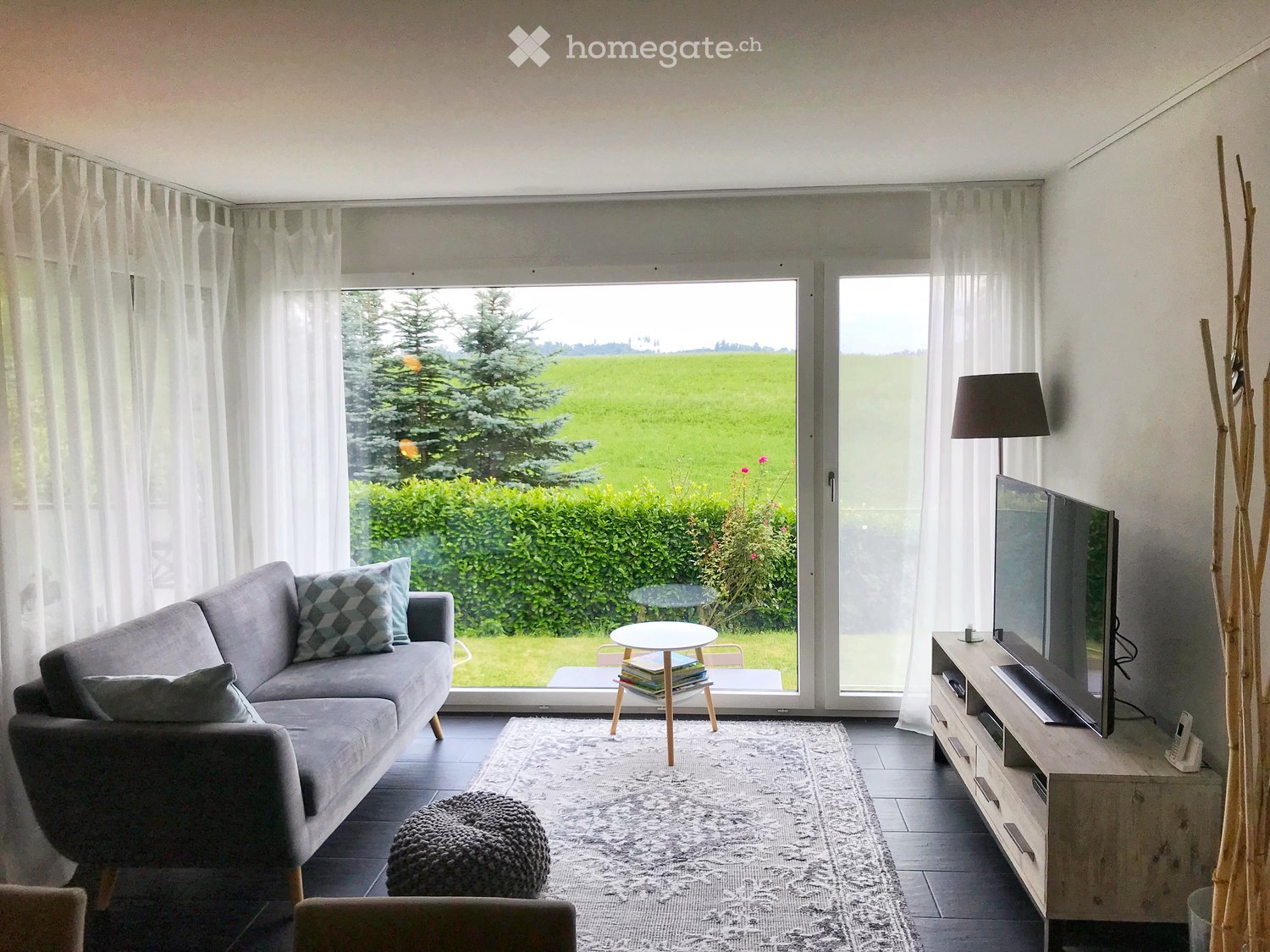 Modern living room with large windows overlooking a green field, featuring a grey sectional sofa, coffee table, ottoman, TV stand, and a small round table with books.