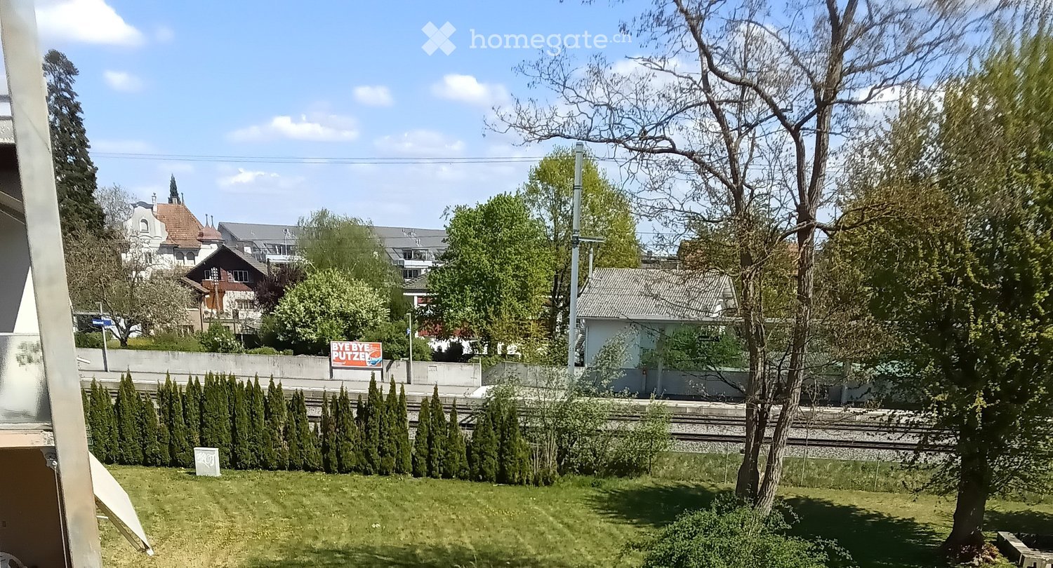 A well-maintained garden with neatly trimmed hedges, a lawn, and trees, with a train track visible in the background and a sign reading "BYEBYE PUTZEL" on a wall.