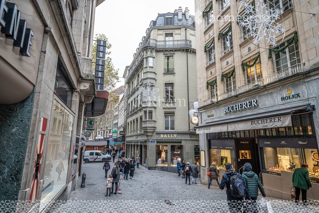 A bustling urban street lined with classic European buildings, featuring luxury shops like Bucherer and Rolex, with pedestrians walking along the cobblestone pavement.