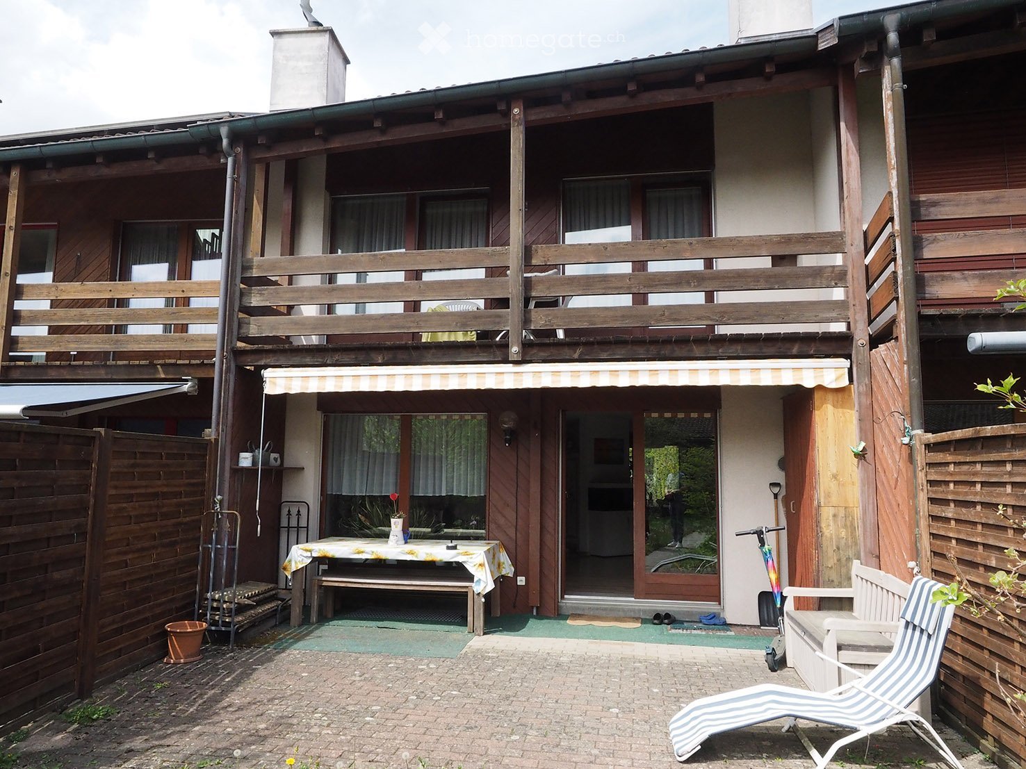 Outdoor patio area with a table, bench, lounge chair, scooter, and a small garden area with a wooden fence and a wooden deck.