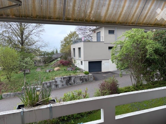 View from a balcony overlooking a modern house with a garage, surrounded by greenery and a stone retaining wall.
