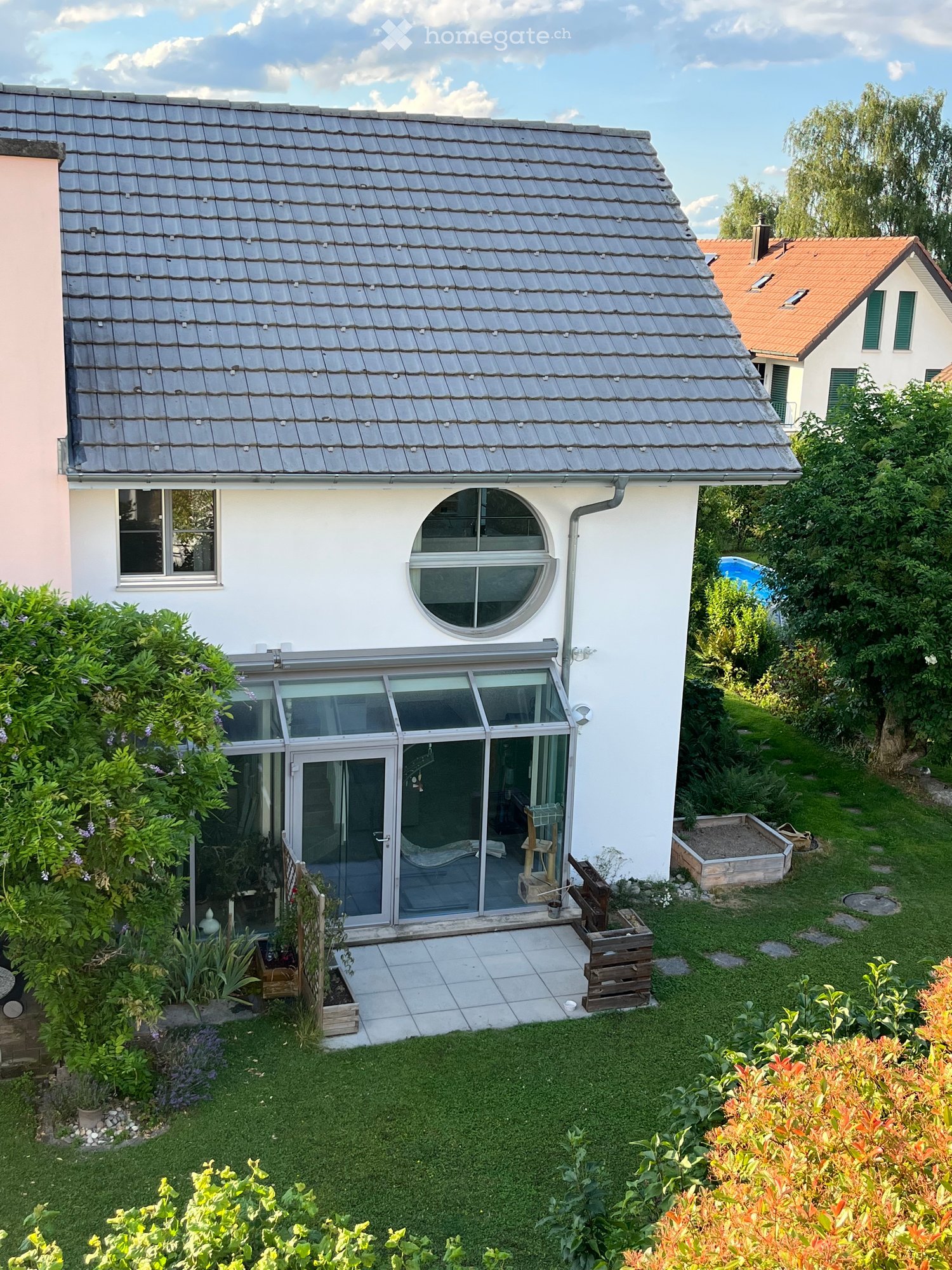 Modern single-story house with a grey tiled roof, white exterior walls, large windows, and a spacious garden with a patio, plants, and a swimming pool.