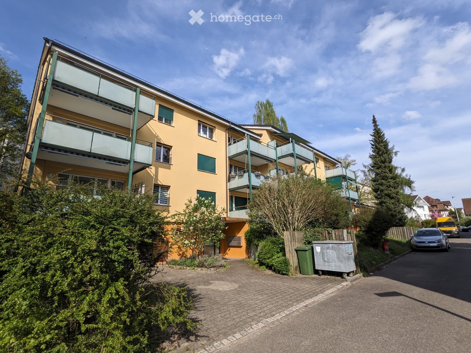 A multi-story residential building with yellow walls, green balconies, and surrounding greenery.