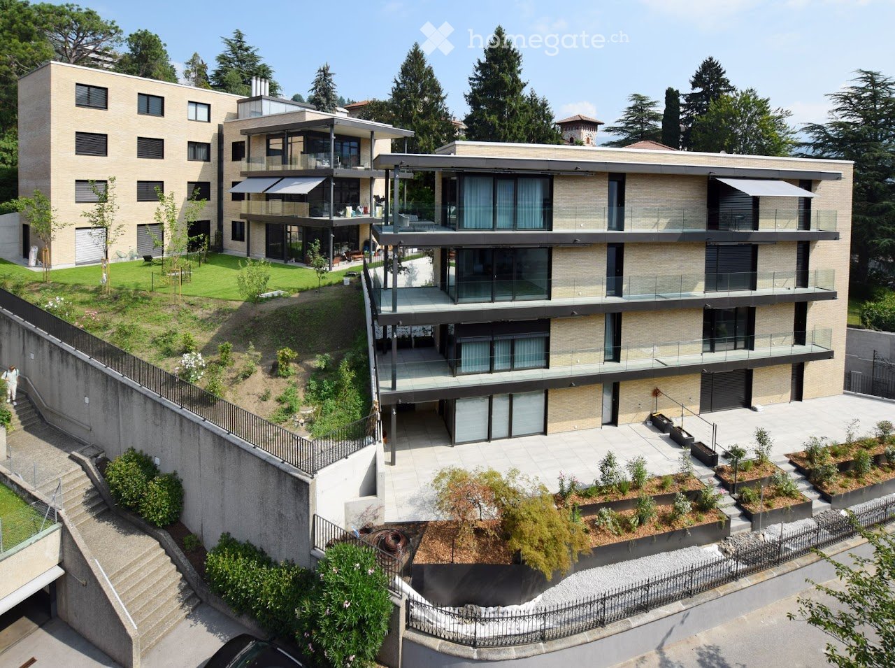 Modern multi-story building with balconies, raised ground floor, and landscaped garden area.