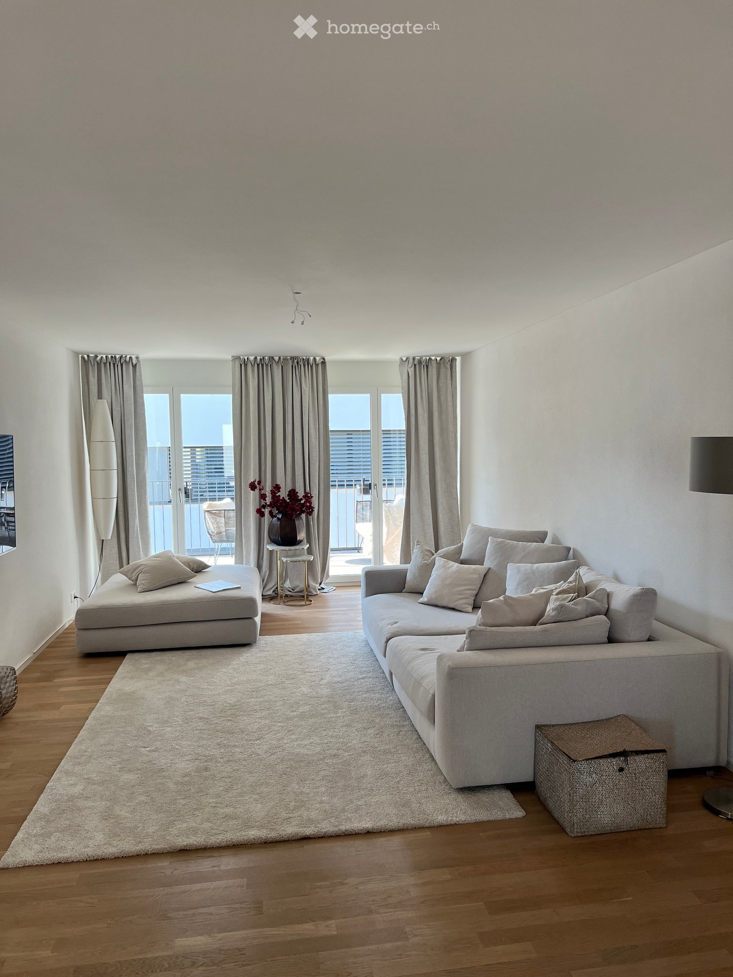 Modern living room with light grey sofa, ottoman, small round table with vase, floor lamp, and large windows leading to a balcony.