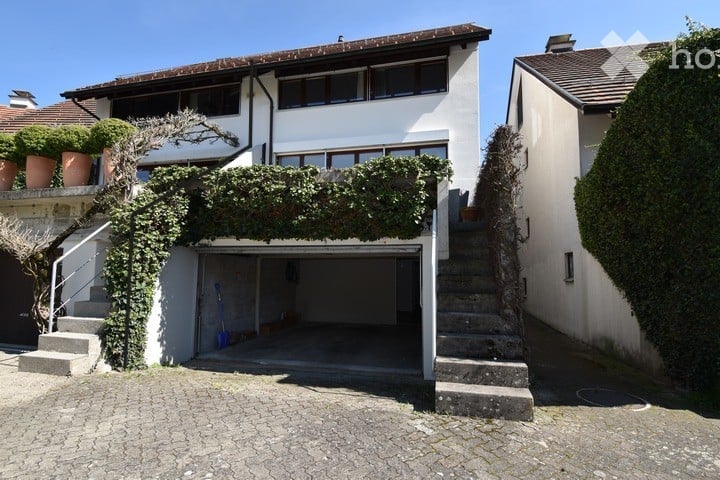 Two-story white building with a garage entrance, surrounded by greenery and potted plants on the upper level.