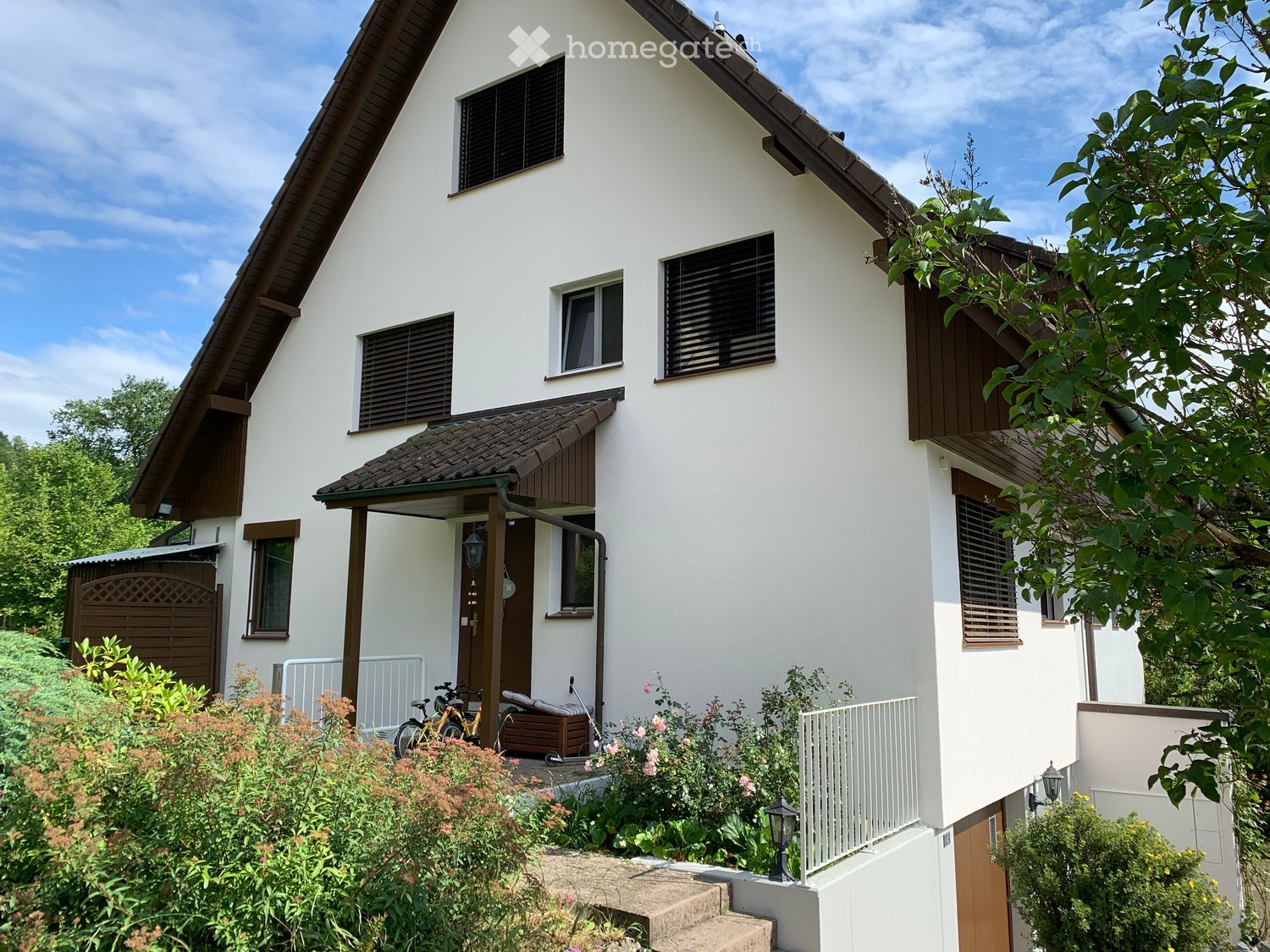 Two-story white house with brown trim, multiple windows with shutters, garage, and small garden area with plants and a bicycle.