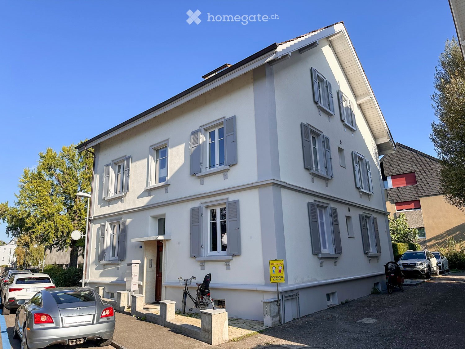 Two-story white building with multiple windows and shutters, parked cars in front, bicycle, and a sign warning about playing children.