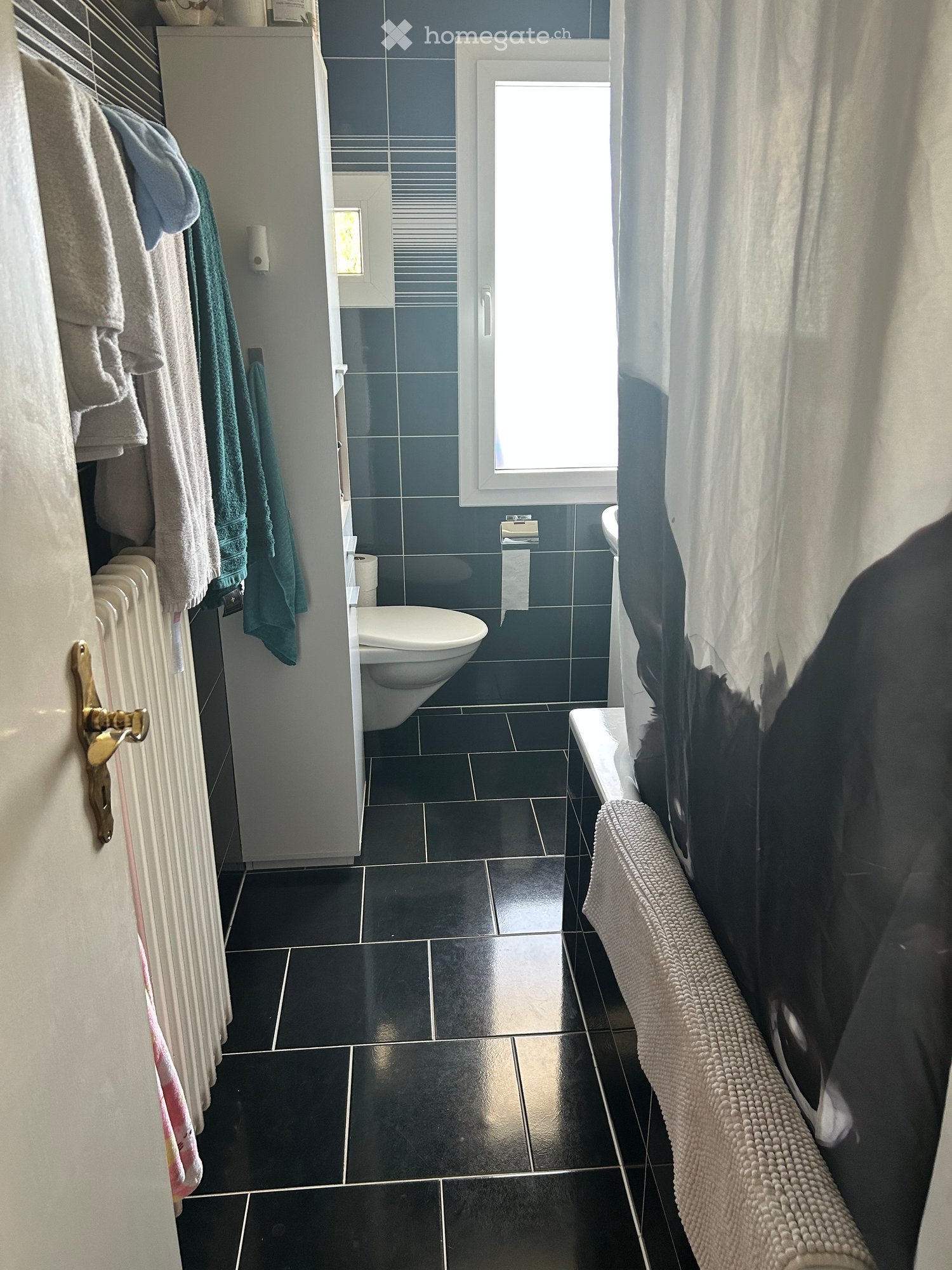 Modern bathroom with dark tiles, a toilet, a radiator, towels, and a window with a curtain.