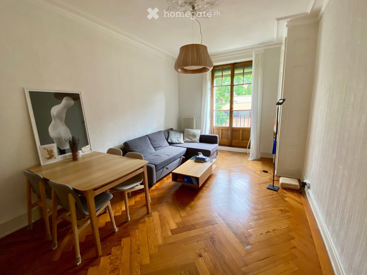 Bright living room with wooden flooring, a grey sectional sofa, a wooden coffee table, a dining table with chairs, and a large mirror on the wall.