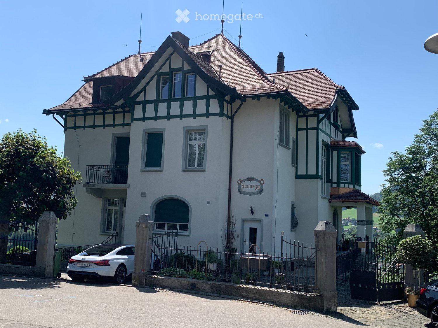 Two-story house with white walls, green trim, red-tiled roof, multiple windows, and a balcony. A white car is parked in front of the house behind a metal fence.