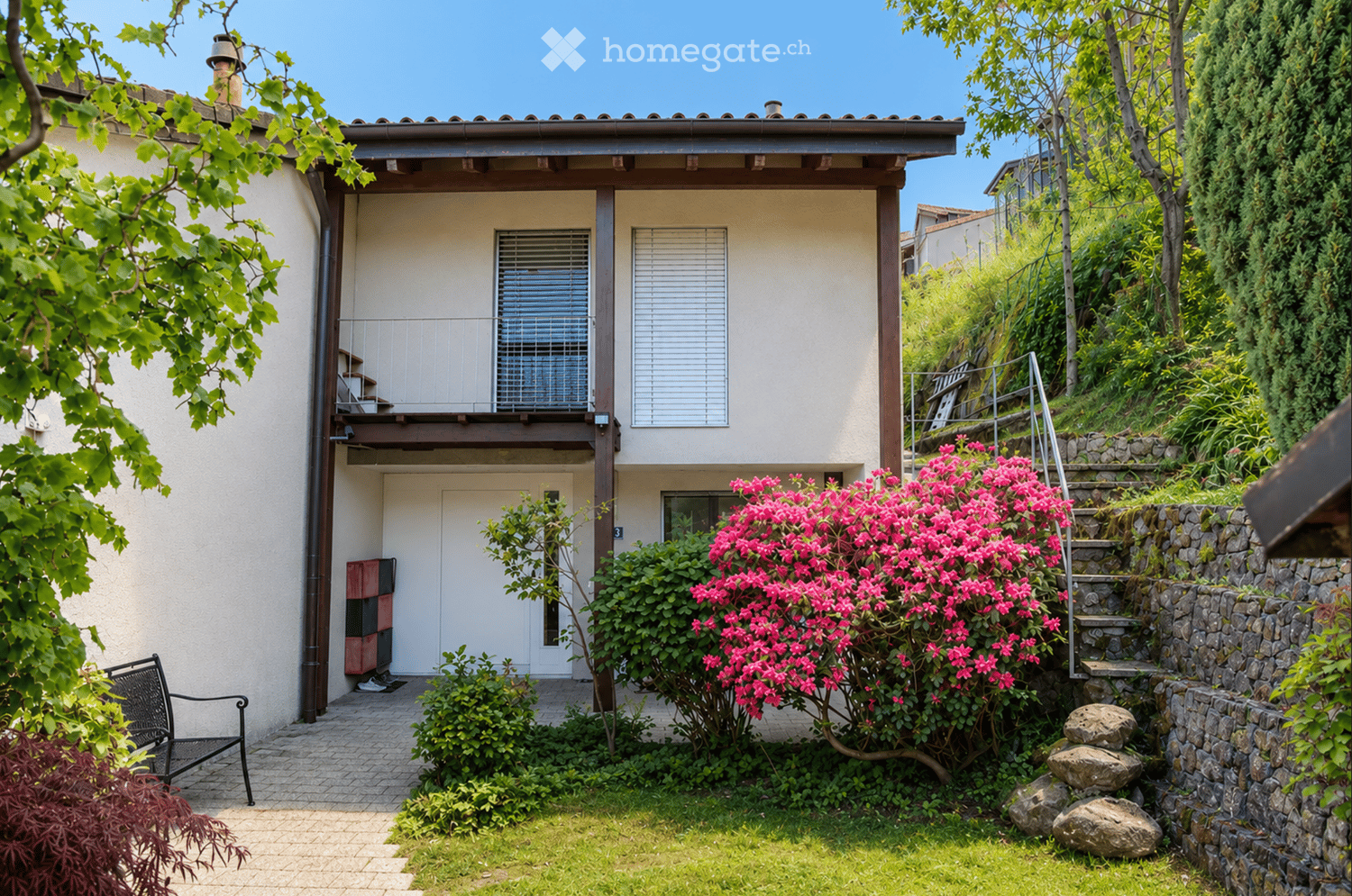 Two-story house with a raised ground floor, beige facade, and a small balcony on the upper level. The entrance is surrounded by a well-maintained garden with vibrant pink flowers, shrubs, and a stone staircase leading up to the house.