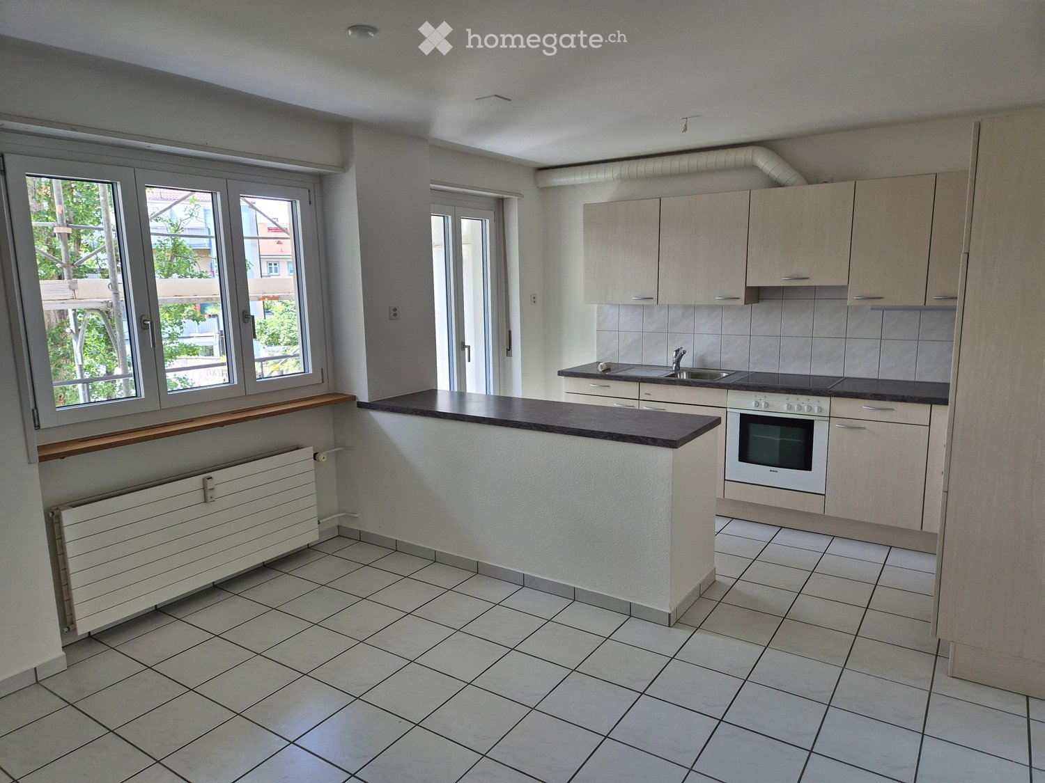 Modern kitchen with light-colored cabinets, a central island, tiled floor, and large windows offering a view of the outdoors.
