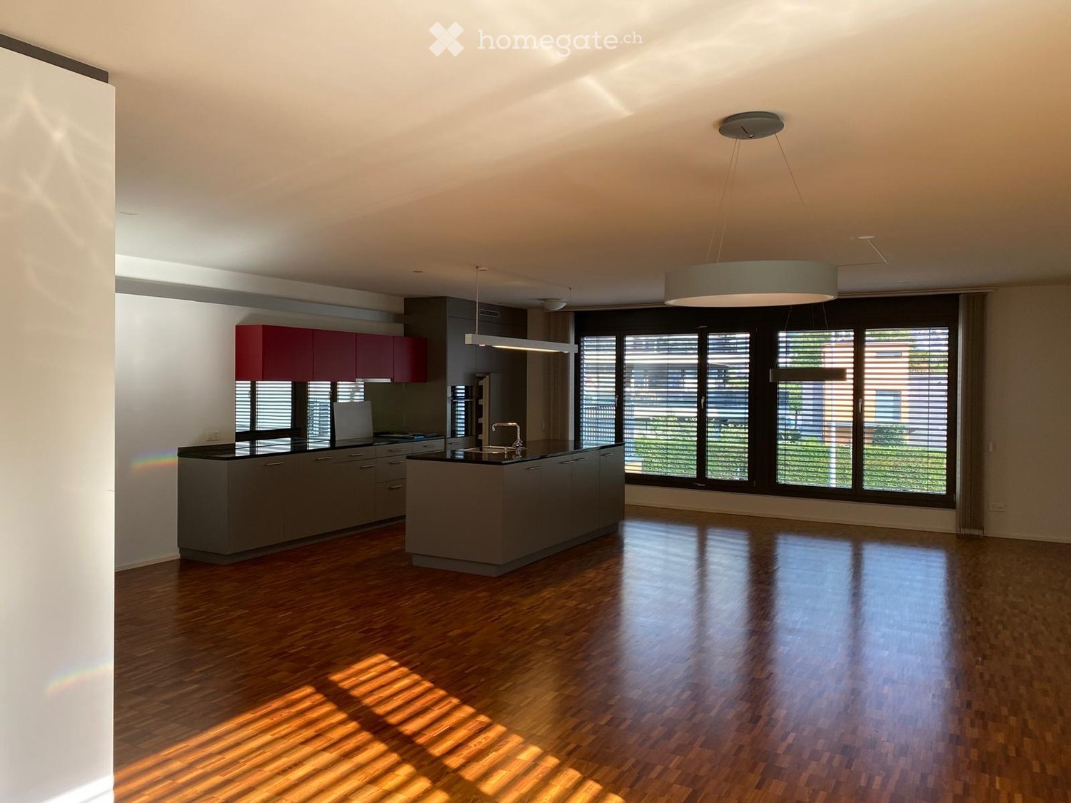 Modern kitchen with a central island, dark grey cabinets, and large windows with blinds. The floor is made of polished wood, and the space is well-lit with pendant lights.