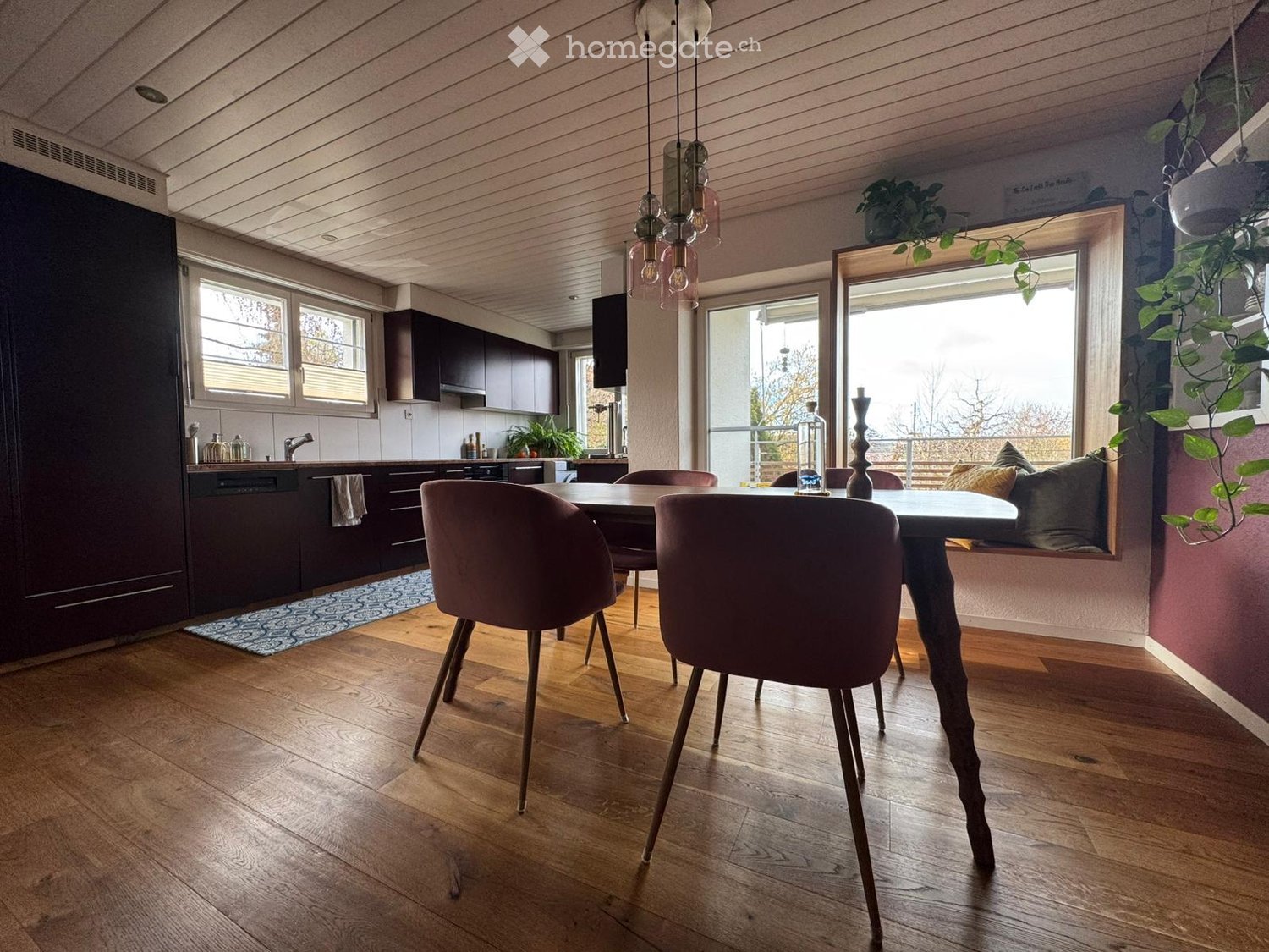 Open-plan dining and kitchen area with wooden flooring, modern kitchen cabinets, a dining table with chairs, pendant lighting, and a window with a view.