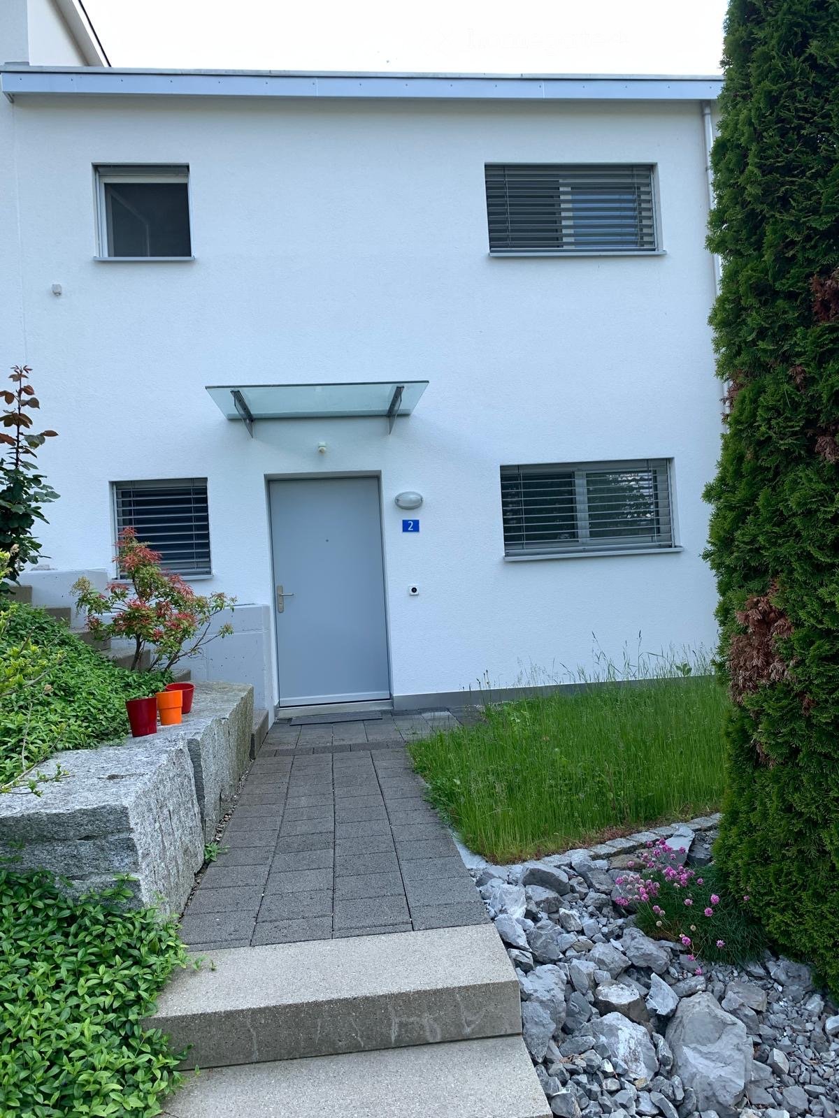 Single-story white house with a grey front door, three windows with blinds, and a small garden area with potted plants and a pathway.